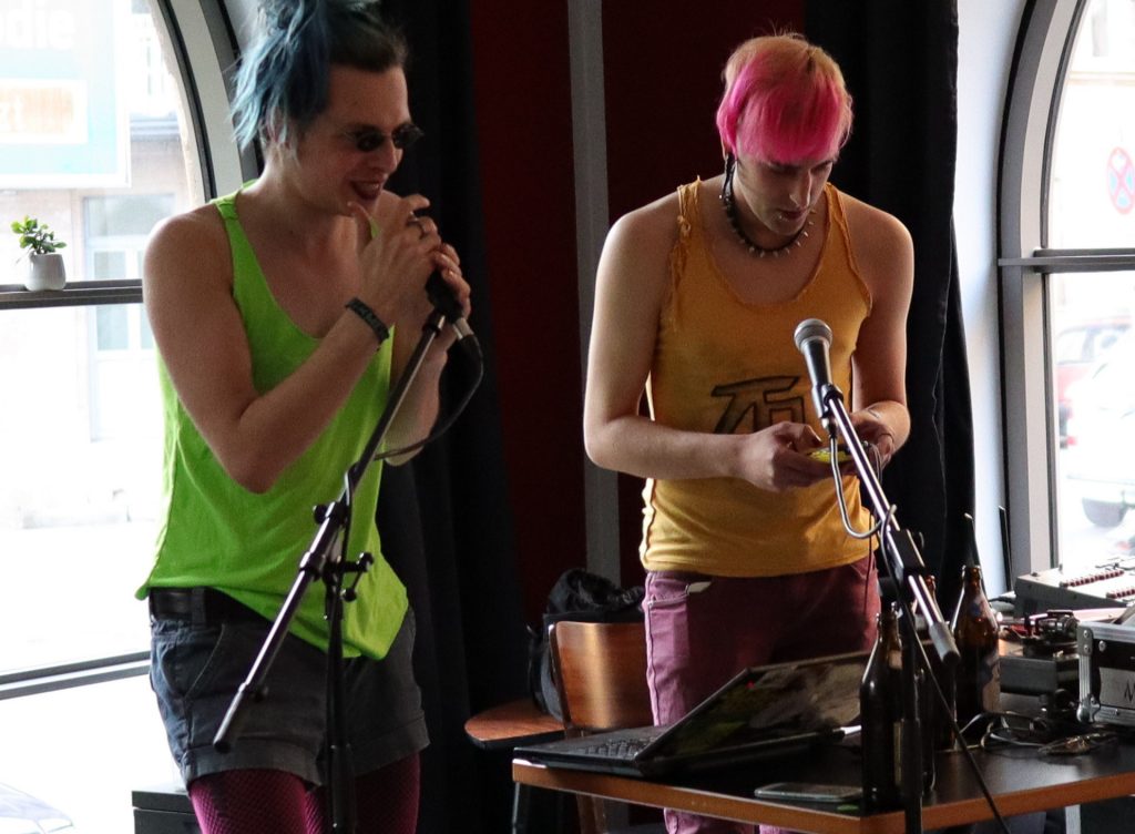 Zwei cyberpunks stehen auf einer Bühne. Einer singt in ein Mikrofon, der andere spielt auf einem Synthesizer.