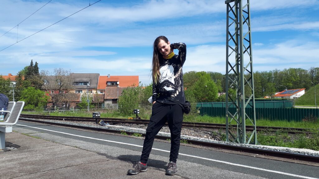 Nami steht an einem Bahnsteig und trägt einen Billie-Eilish-Pullover mit der Aufschrift "Happier than ever".