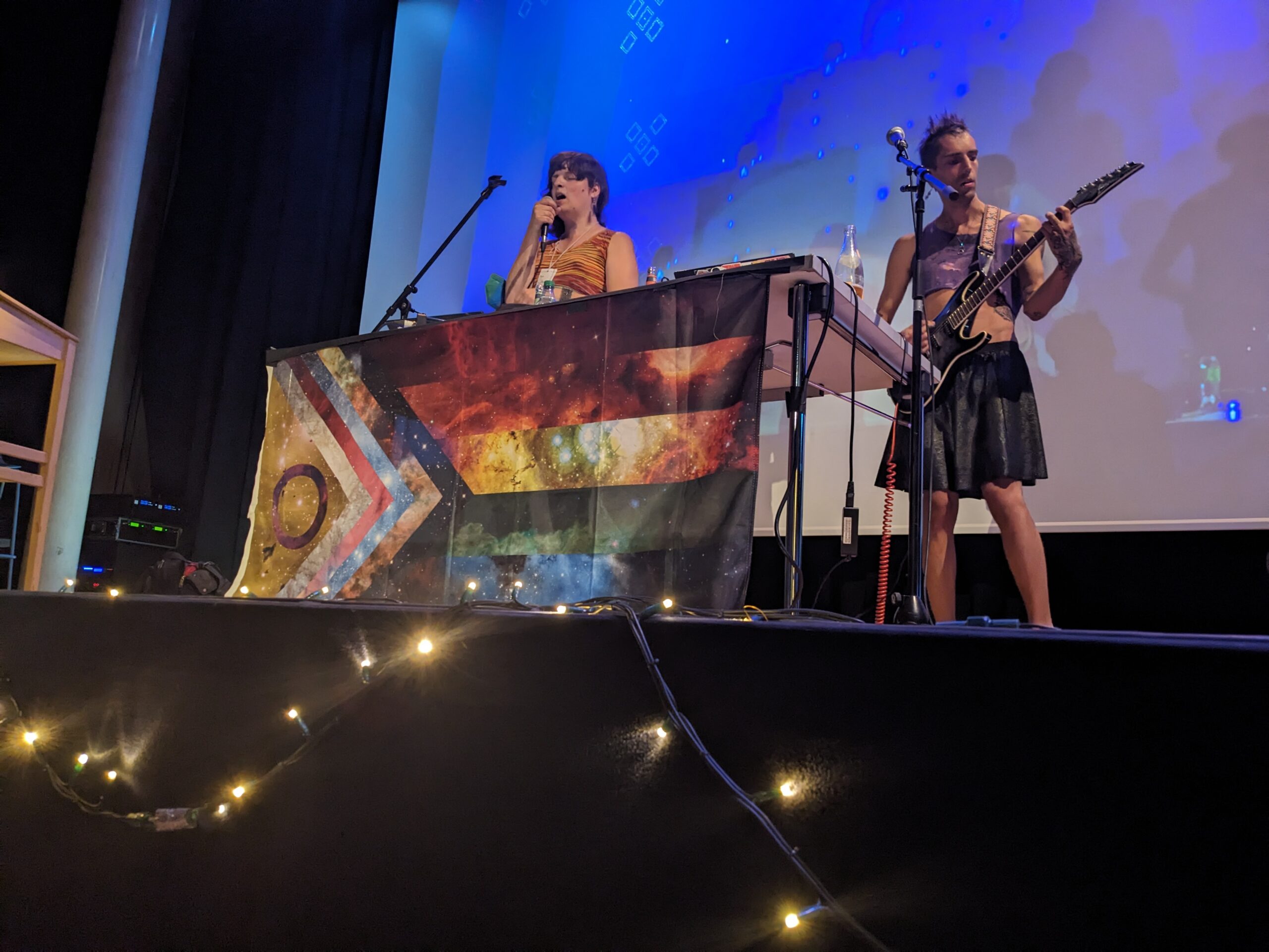 Nami & Phant auf einer Bühne, vor ihrem Tisch hängt eine riesige progressive-Pride-Flagge, hinter ihnen ist der Hintergrund blau mit repetetiven Sillouetten der beiden.