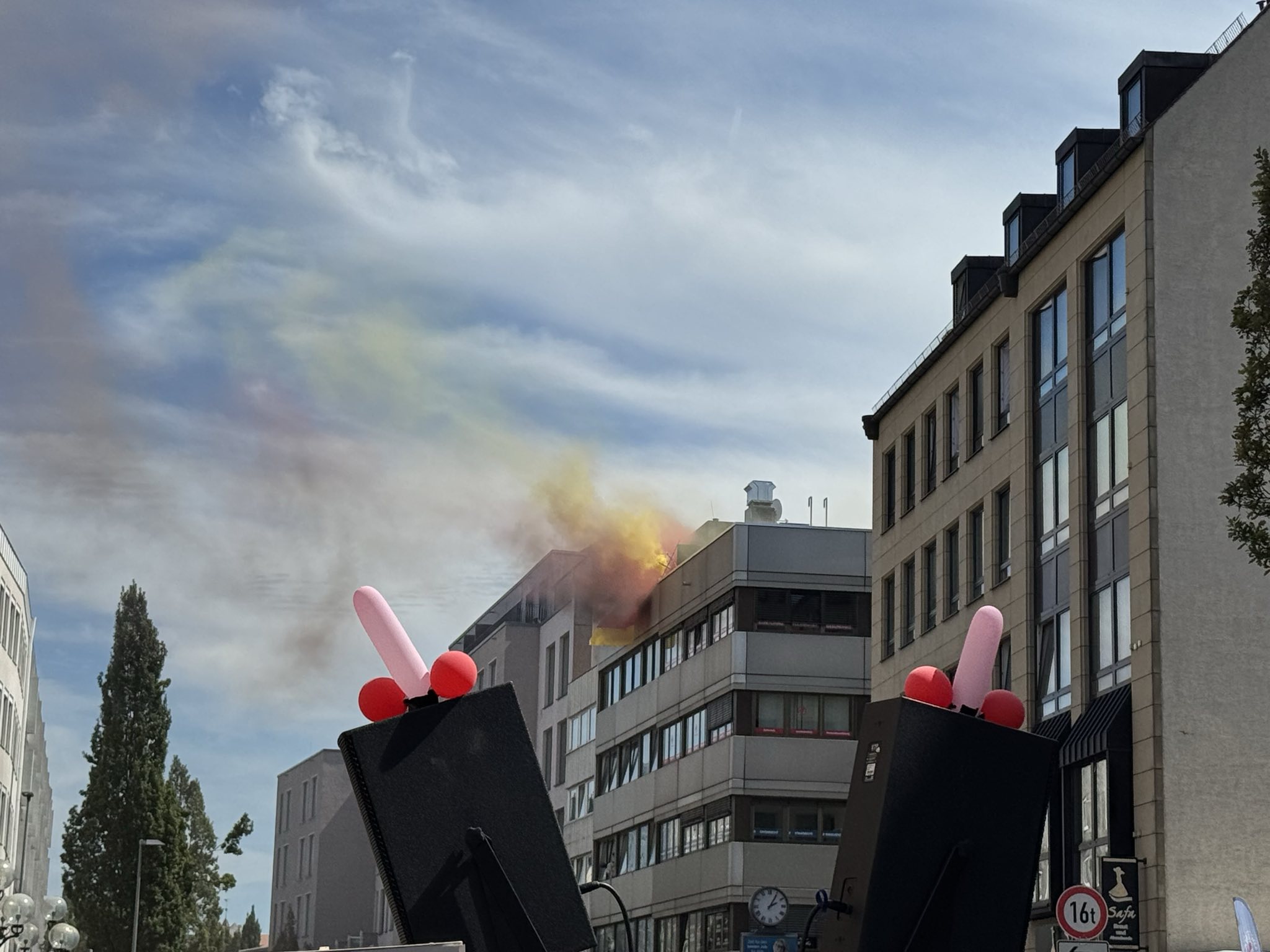 Zwei große Boxen, auf ihnen sind Penis-Luftballons. Im Hintergrund ist ein Gebäude zu sehen, auf dessen Dach eine bunte Rauchfackel gezündet wurde.